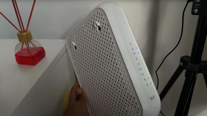 Router WiFi blanco con ventilación y LEDs de estado, sostenido con la mano sobre una mesa blanca.