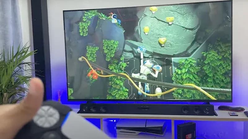 Mando PS5 DualSense frente a TV con Astro Bot en PS5 Slim sobre mueble con retroiluminación azul