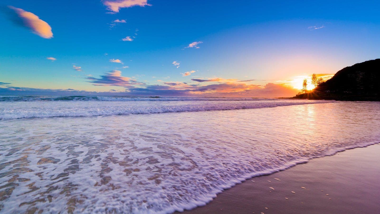 Atardecer en playa con olas espumosas, cielo azul y naranja dorado, árboles y acantilado al fondo.