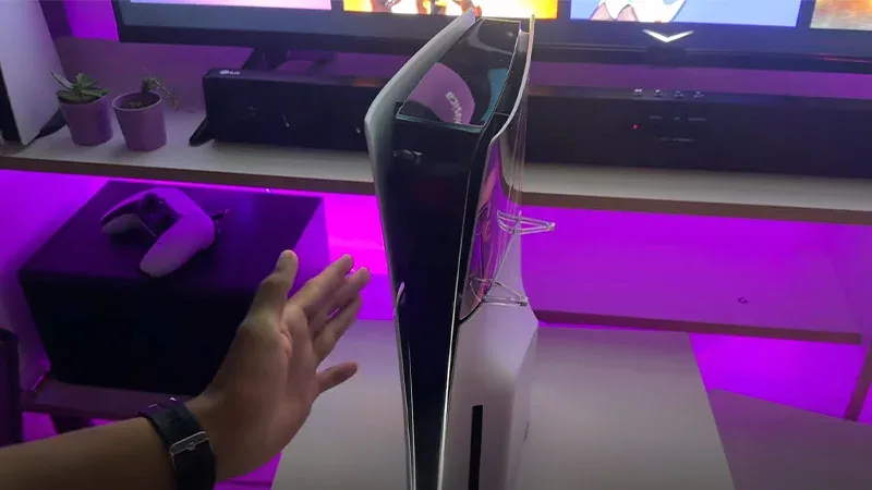 PS5 Slim en posición vertical sobre escritorio con iluminación RGB morada y mano señalando la consola