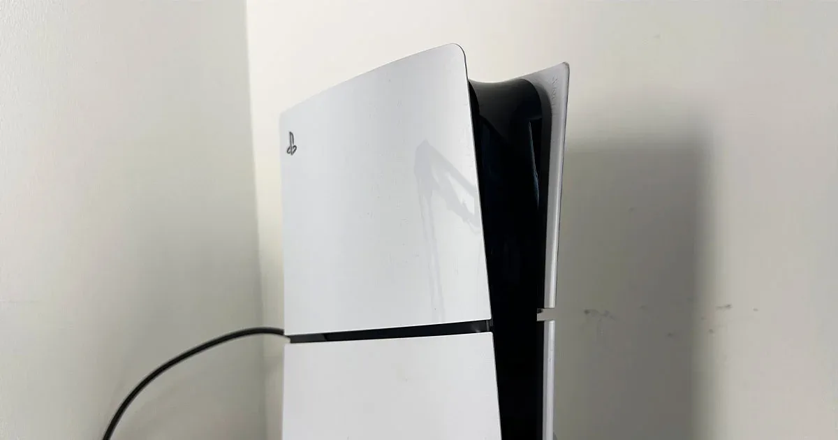 Consola PS5 Slim de Sony en blanco y negro colocada verticalmente junto a una pared con cable conectado.
