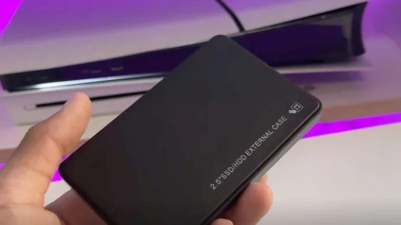 Mano sosteniendo caja externa 2.5" SSD/HDD frente a una PS5, ideal para ampliar almacenamiento de la consola.
