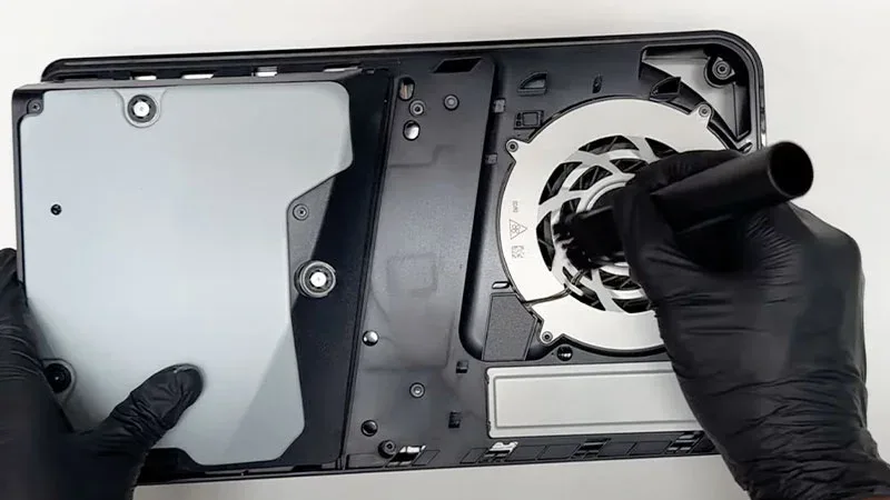 Limpieza del ventilador interior de una PS5 Slim con cepillo, usando guantes negros de látex.