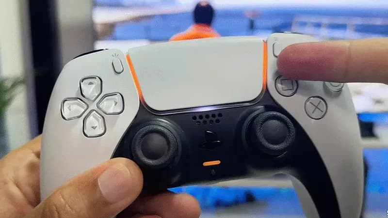 Mando DualSense de PS5 con luz naranja en mano, mostrando botones táctiles, cruceta y joysticks