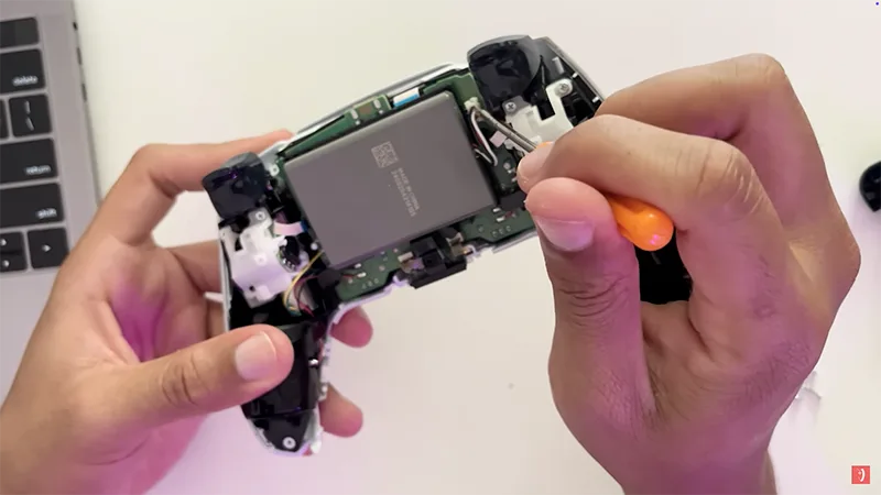Desmontaje interior del mando DualSense de PS5 con destornillador naranja mostrando batería y placa base