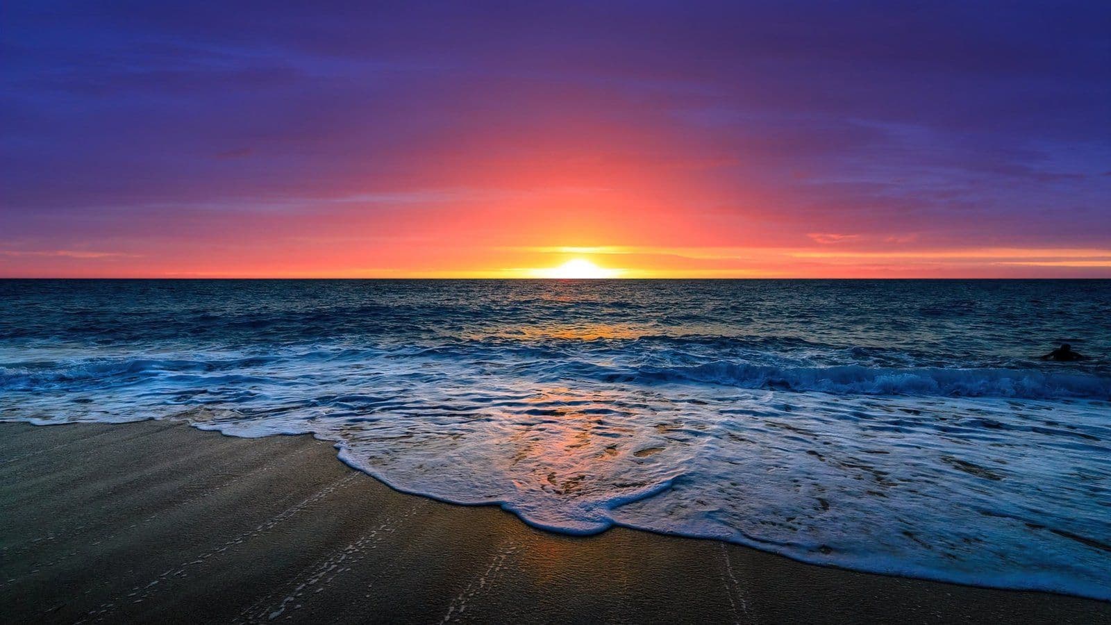 Atardecer en la playa con cielo púrpura y naranja, olas suaves y reflejo dorado del sol en la arena húmeda.