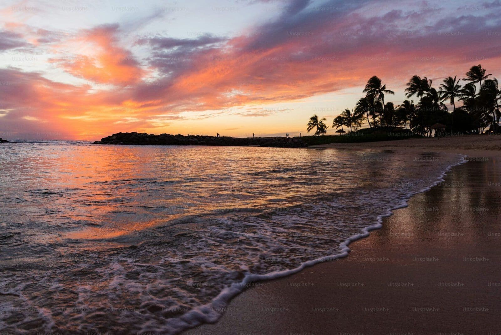 Atardecer tropical en playa con palmeras, cielo naranja y olas suaves reflejando colores cálidos del ocaso.